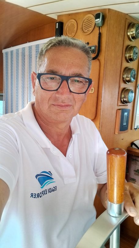 Man in a white polo shirt, wearing glasses, posing on a boat with nautical equipment.