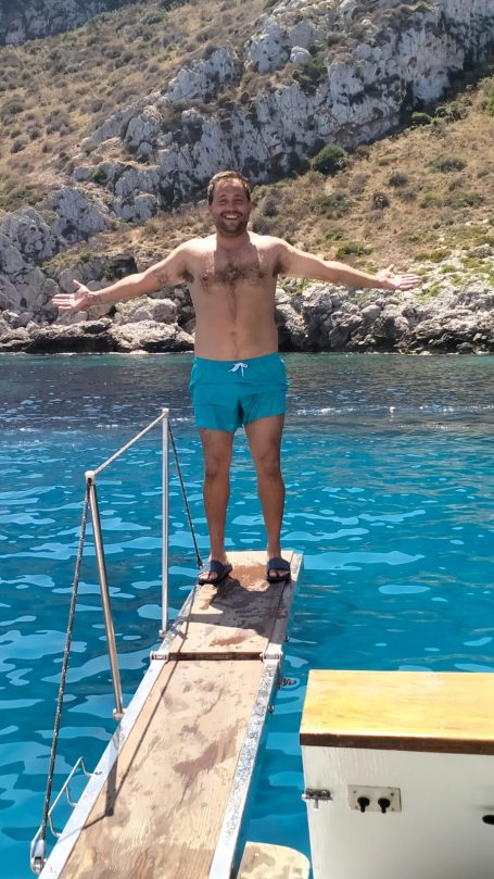 Uomo in costume da bagno blu che si trova su un molo, con le braccia aperte in un mare turchese.